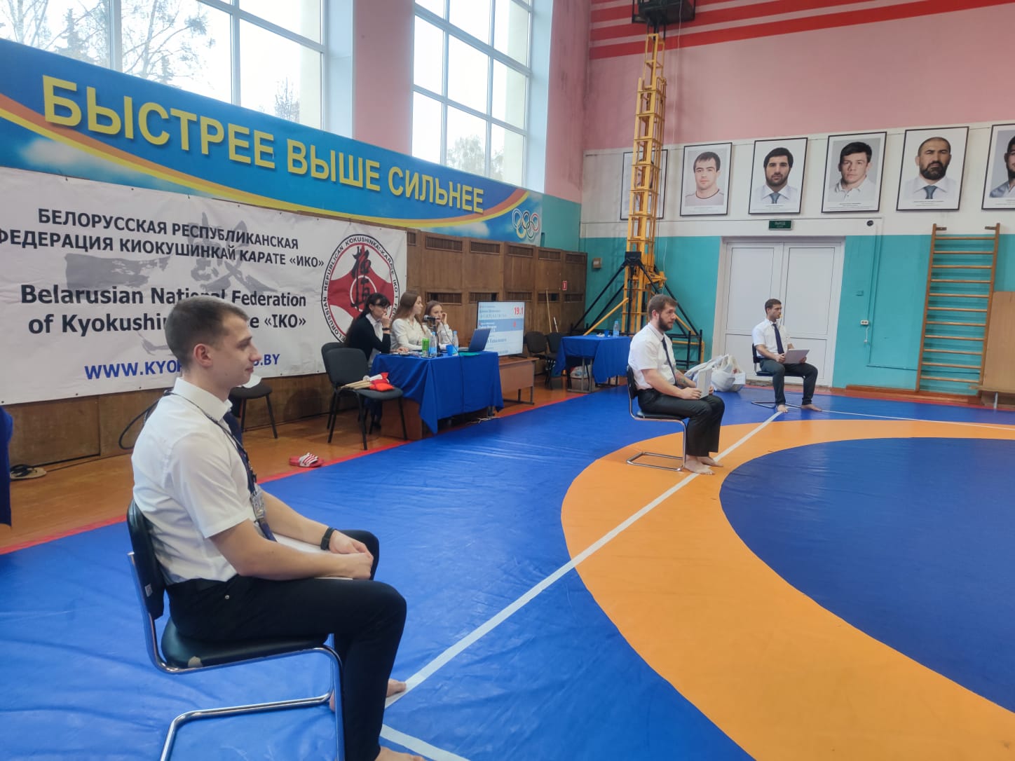 Судьялардың артында „ИКО“ Каратэ Федерациясы баннері: „Жылдам. Жоғары. Күшті“ Belarus Open Cup 2023 ашық халықаралық турнирі залындағы татамидағы төрестер тобы және хатшылық