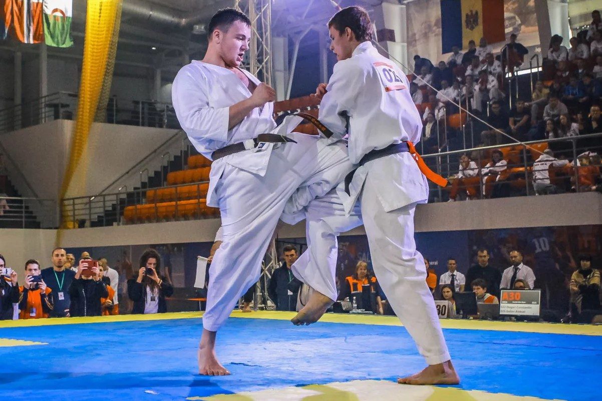 Am doi luptători cu centură neagră; în fundal — drapelul Kazahstanului și tabla „130“ Duel kumite la Al 17-lea campionat european deschis de karate: lovitura cu piciorul în sus, public și fotografi în arenă
