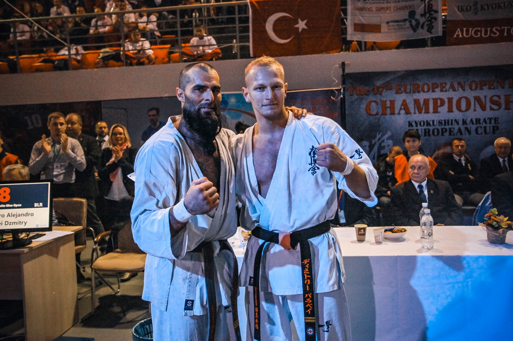 Pe banner — „Kyokushin Karate European Cup“; pe monitor — codul țării BLR Doi karateka pe scenă la Al 17-lea campionat european deschis de karate, cu bannerul evenimentului în fundal