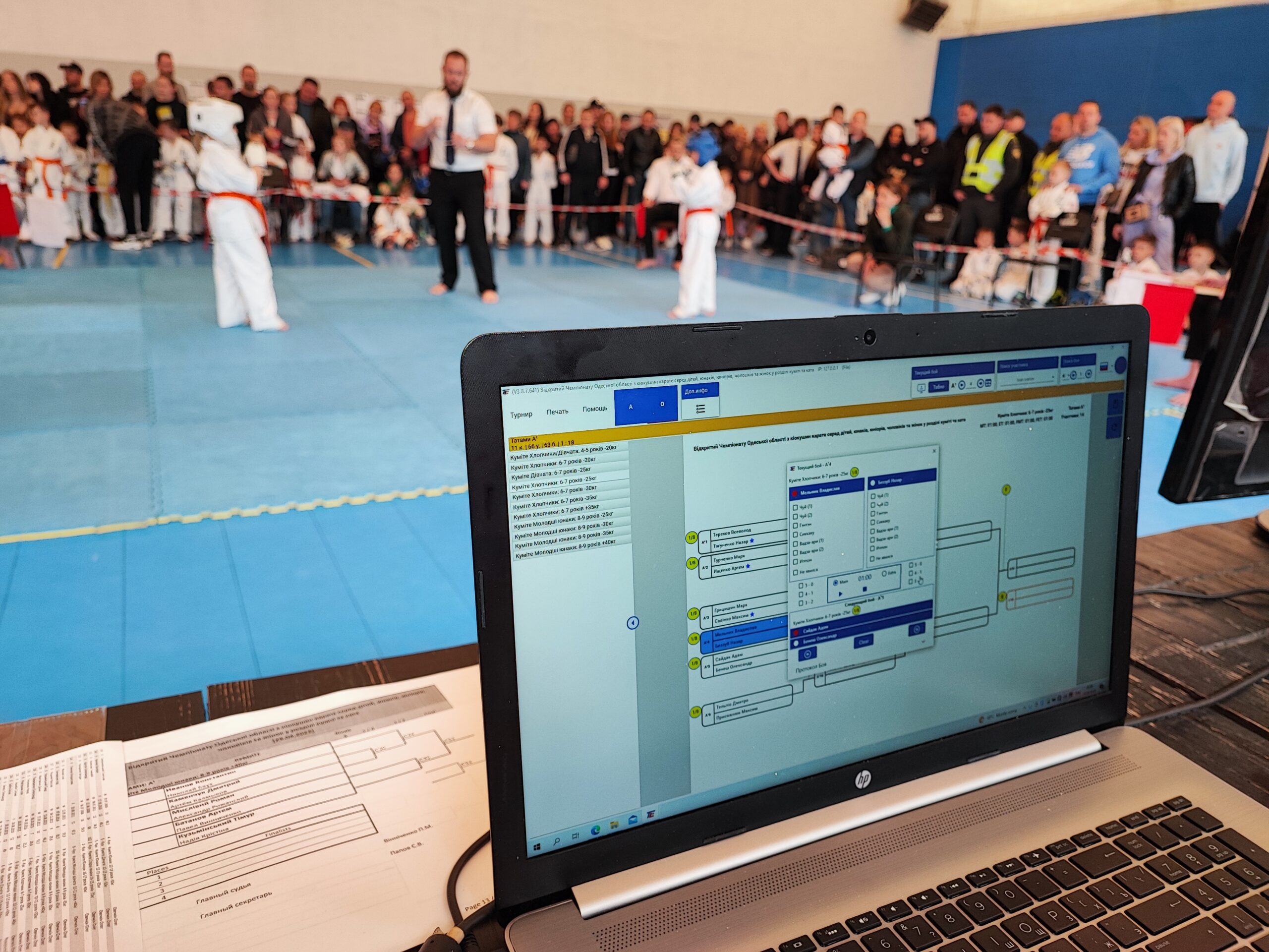 «Одесса облысының кіокушин карате бойынша чемпионаты» бағдарламасы, «А14» торы, 29.04.2023 Алдыңғы планда турнир кестесі бар ноутбук, артында Одесса облысының чемпионатында балалар каратистерінің кездесуі