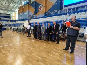 Қара футболка киген адам қызыл кітаптан оқид; артында — костюм киген делегаттар Спорт залында ашылу рәсімі алдындағы Единое Каратэ киокушин-каратэ турнирінің қазылары мен ұйымдастырушылары