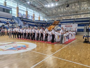 Қызыл кілемде ақ кимоно киген балалар мен ересектер; қызыл жұлдызды логотип; кубок Ақ кимоно киген қатысушылар қызыл татами үстінде Бірлік Каратэ жарысы ашылу рәсімінде