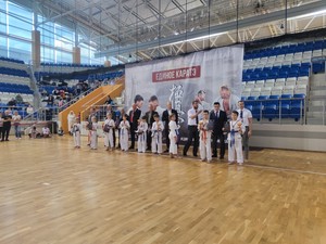 Балалар мен ересектер подиумда; алыста қызғылт сары саты және қызыл жұлдызды баннер Қара белдік иелері және ұйымдастырушылар Бірлік Каратэ баннері алдында топтық суретте