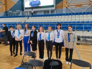 Ақ көйлек пен көк галстук киген волонтерлер; костюм киген ер адам қолымен сөйлейді Кёкүшин-каратэ Бірлік Каратэ жарысы судьялары мен қатысушылары спорт залында